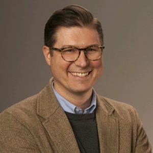 MCN Portrait of Michael Naughton in a brown blazer, smiling confidently.